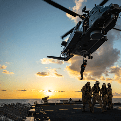 Military personnel on ship with helicopter flying above and soldier dropping being lowered onto deck